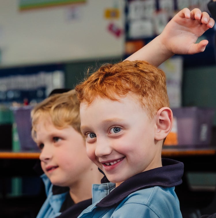 A close up of two young boys smiling. The boy in the foreground is raising his hand.