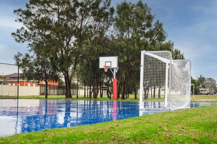 A bright blue outdoor basketball court after fresh rain. A futsal net is positioned to the side of the court and trees can be seen in the distance.