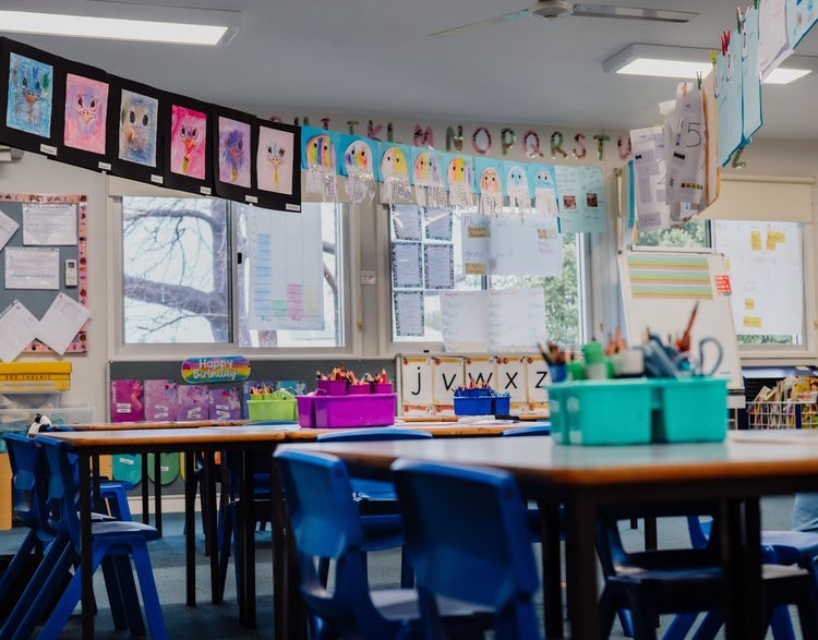 An interior photo of a bright and vibrant classroom showing student desks, chairs and colourful pencil tins. Windows let in light and colourful student artwork can bee seen hanging on walls and from the ceiling. A portable whiteboard can be seen in the distance with learning aids displayed.