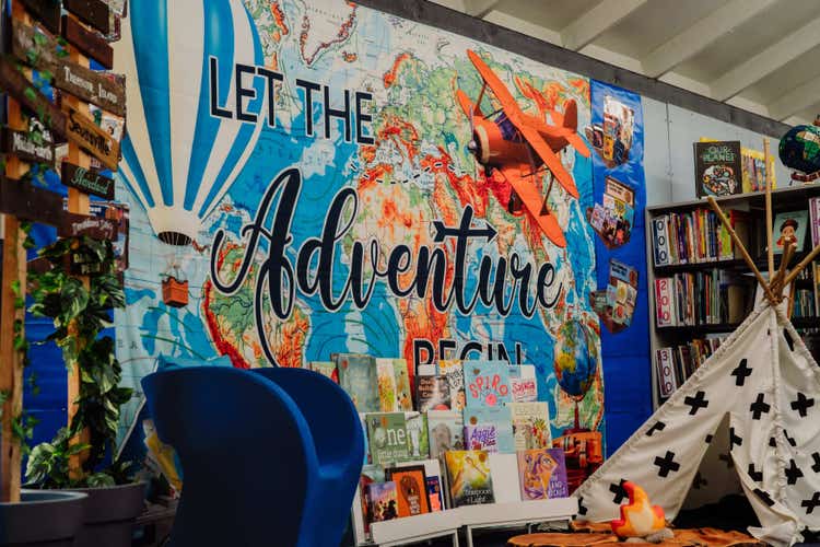 A vibrant library display featuring a large world map mural with the words “Let the Adventure Begin.” The display includes a model airplane, a hot air balloon, a cozy reading area with a small teepee and pretend campfire, and shelves filled with books that invite students to explore and read.