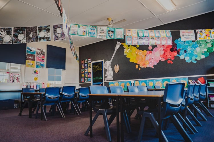 An interior photograph of an art classroom showing student tables and chairs in two large groups. A variety of bright and colourful student artwork is displayed on the walls and hanging from the ceiling.